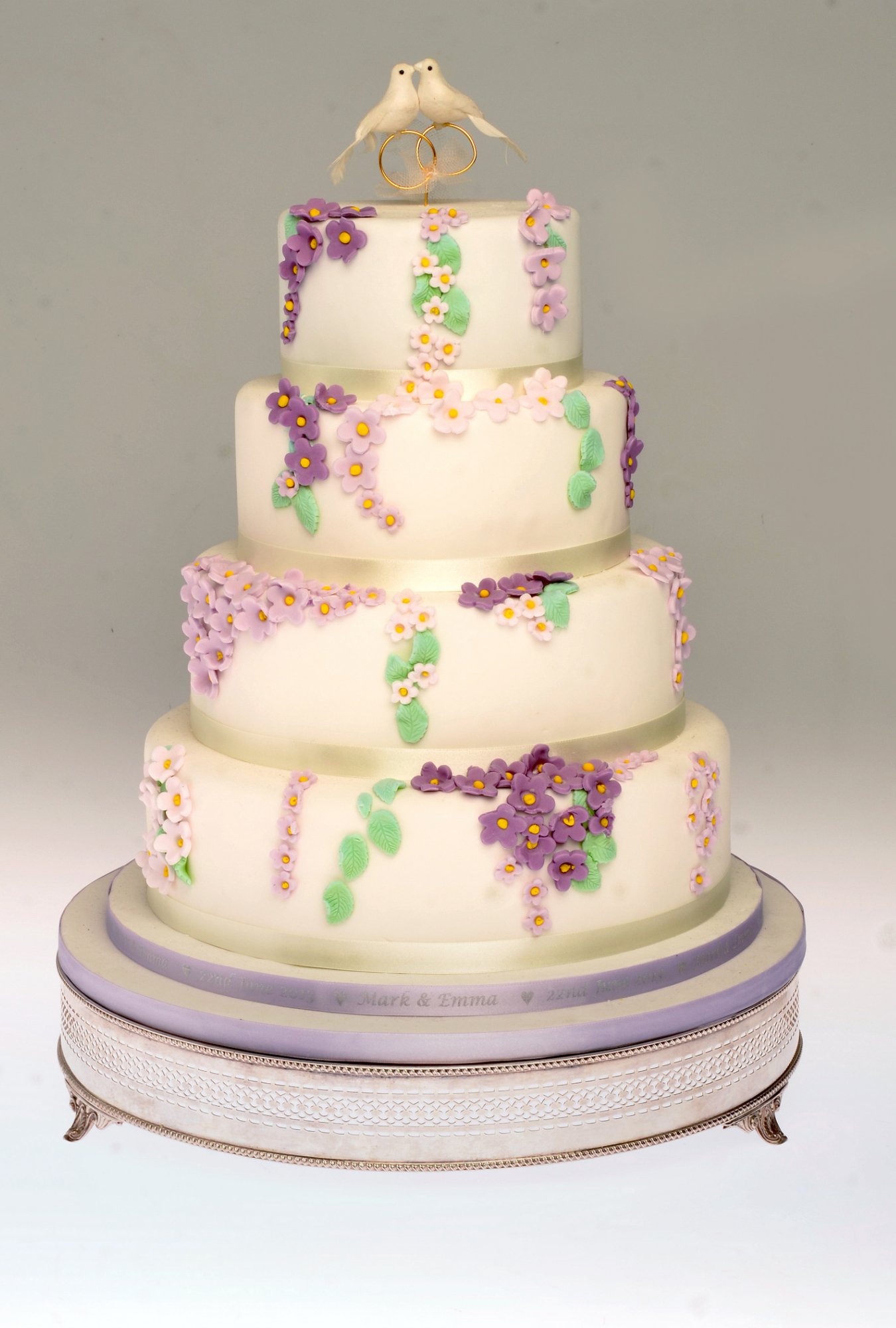 Four tier cream wedding cake decorated with purple and green flowers and topped with two doves over interlocking wedding rings