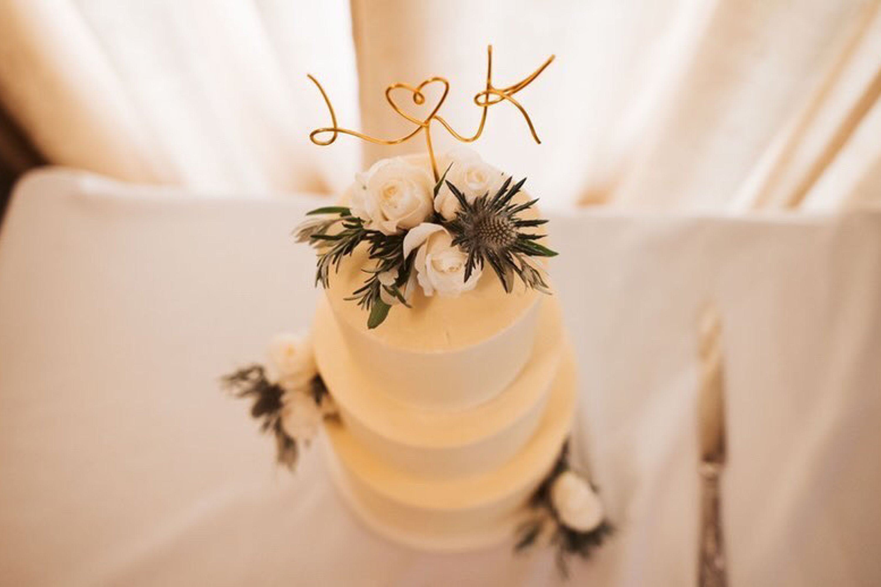 A three tiered cream wedding cake topped with white flowers, pine cones and gold wire initials