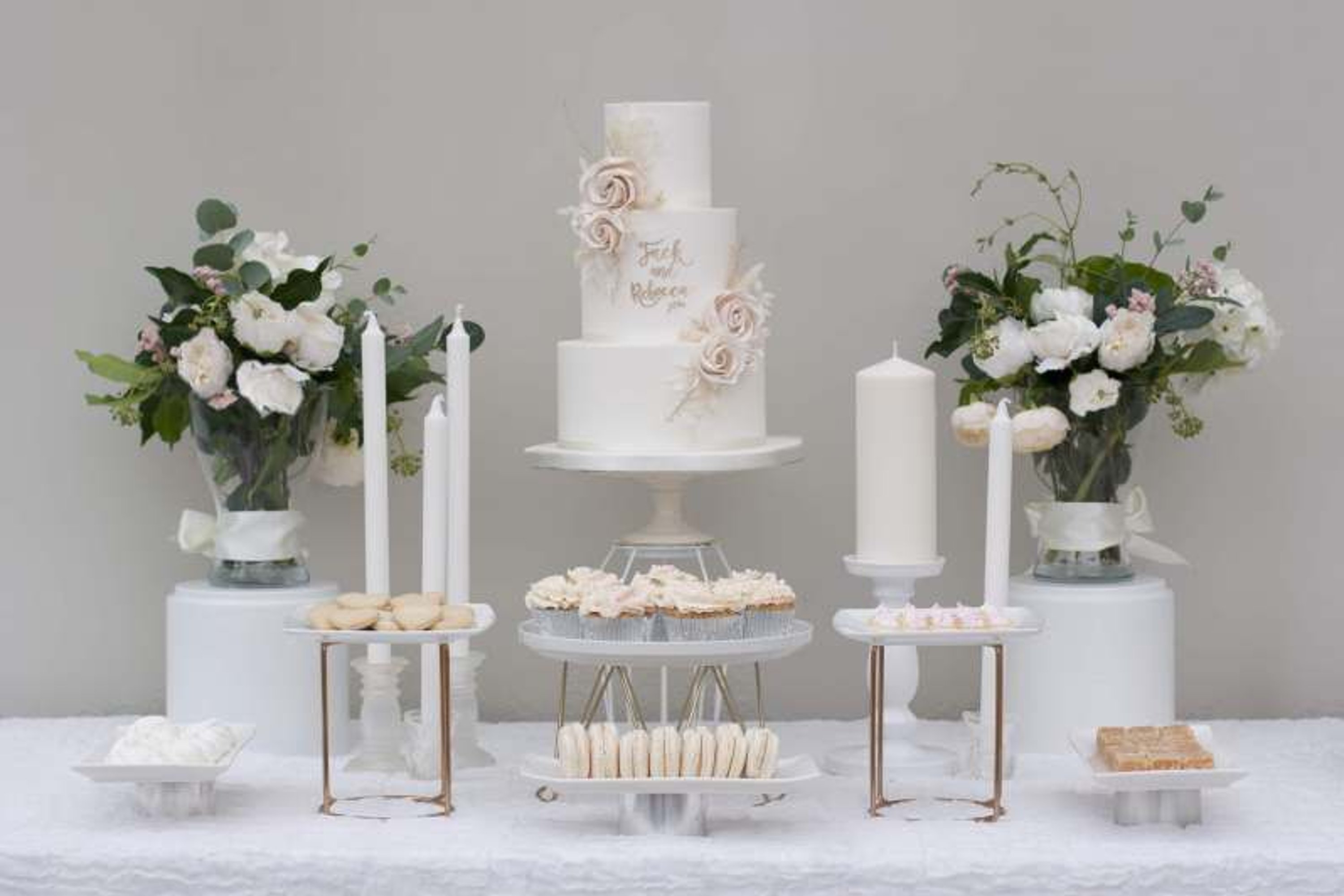 Three tier white wedding cake with beige flowers and white tapered candles on a cream buffet table