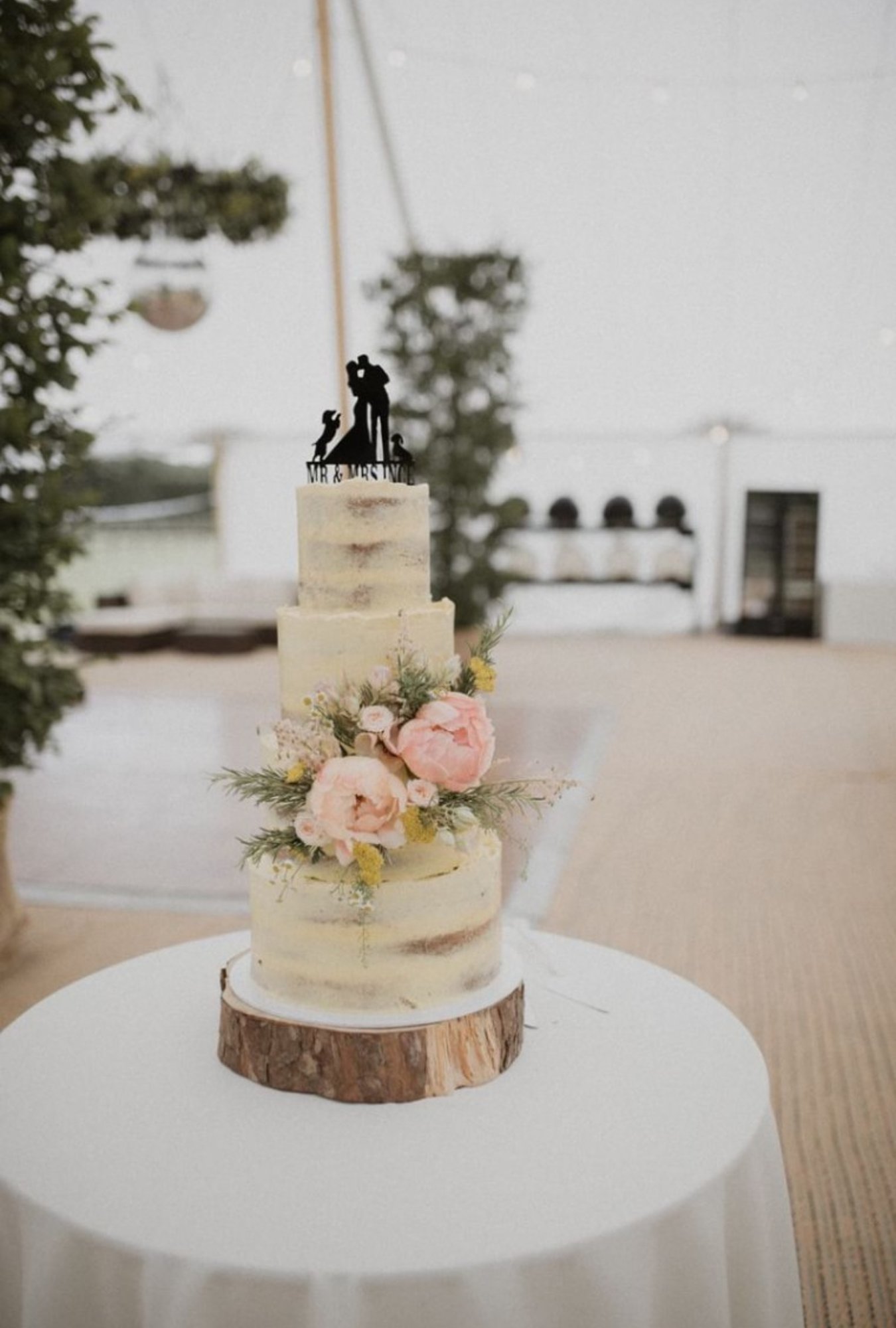 Three tier semi-naked wedding cake with pink flowers topped with a silhouette cake topper of a couple and two dogs