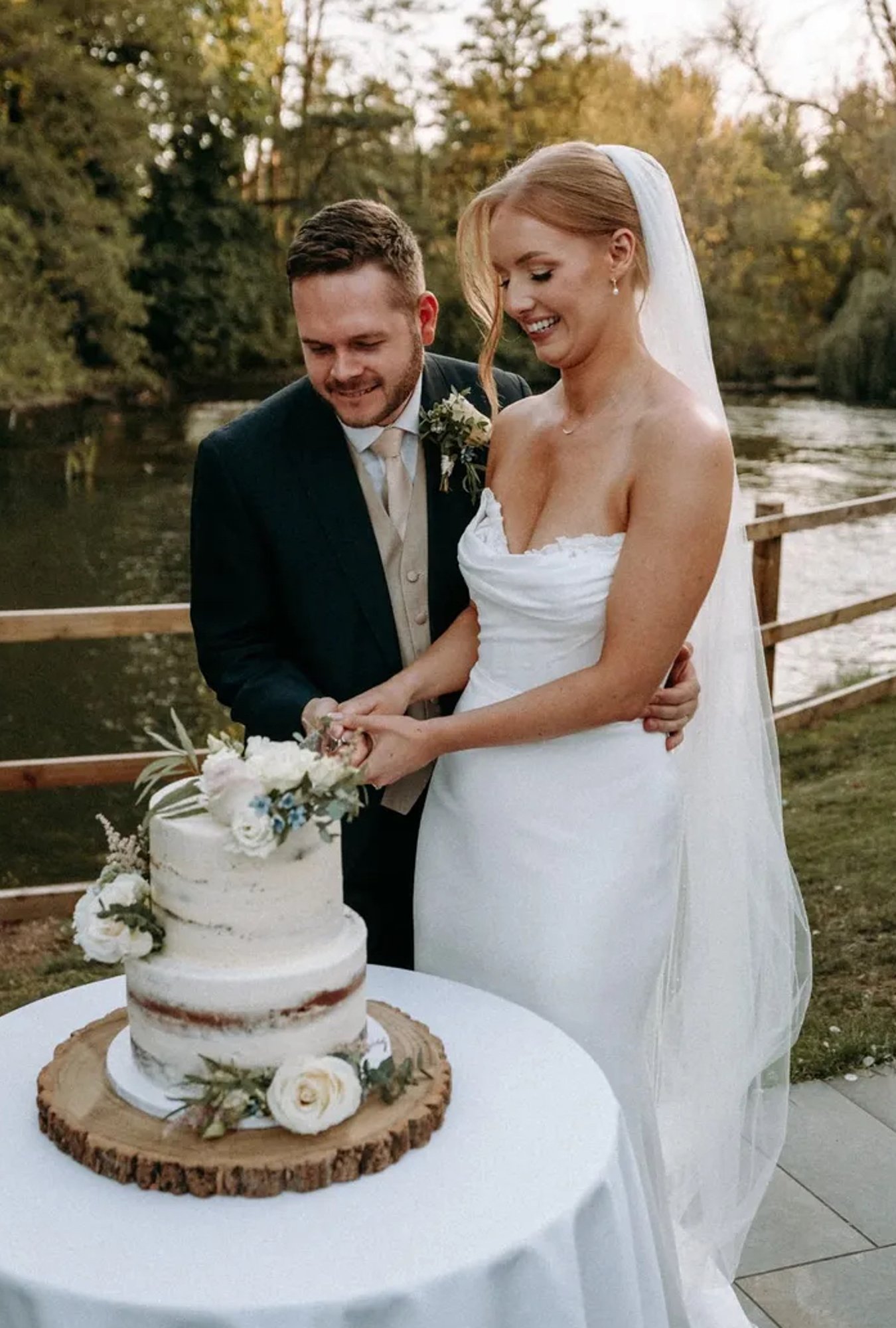 A bride and groom slicing their two tier semi-naked wedding cake outside in front of a lake