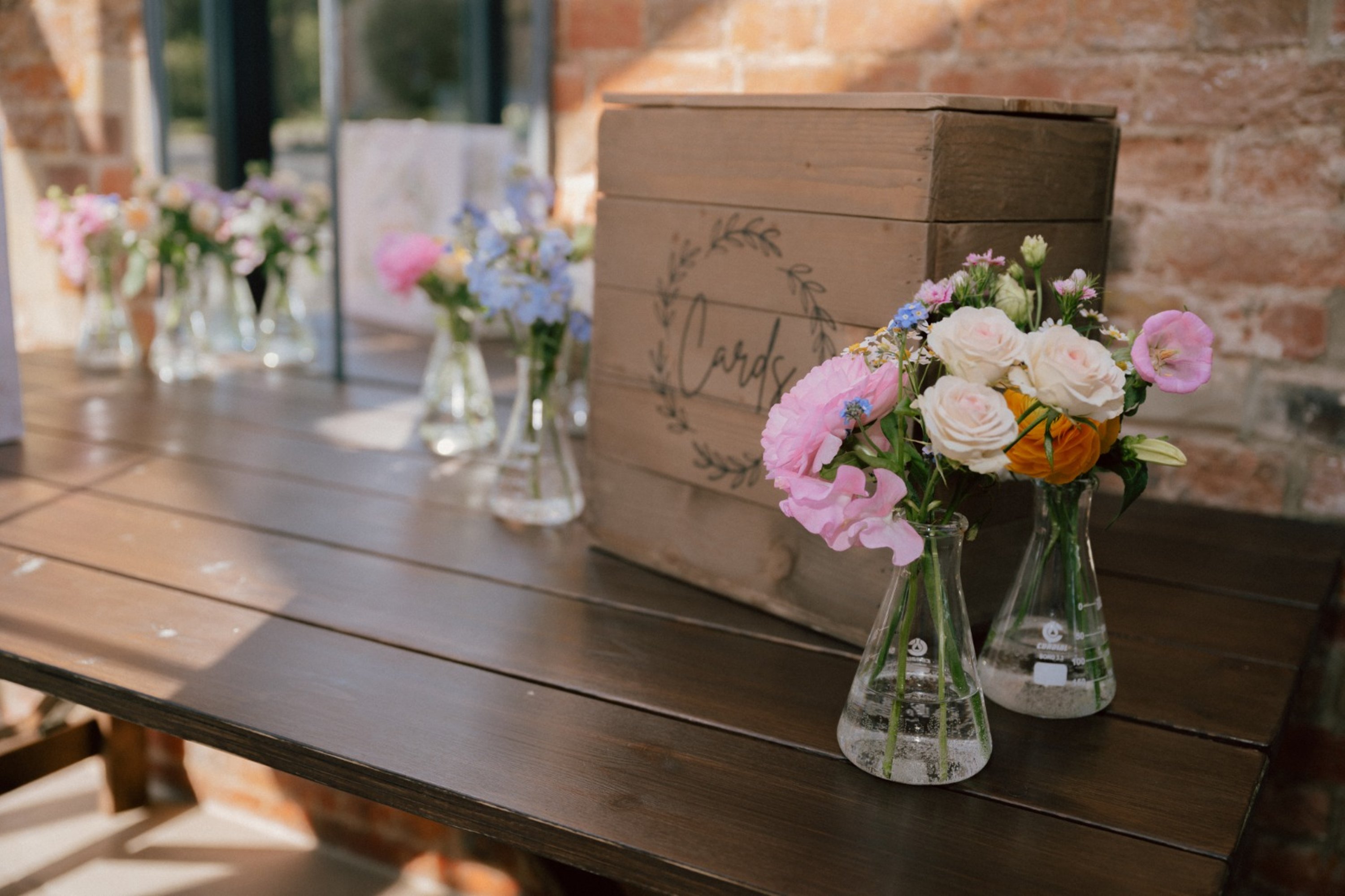cluster of glass vases holding small arrangements of colourful flowers next to a card box on a bench