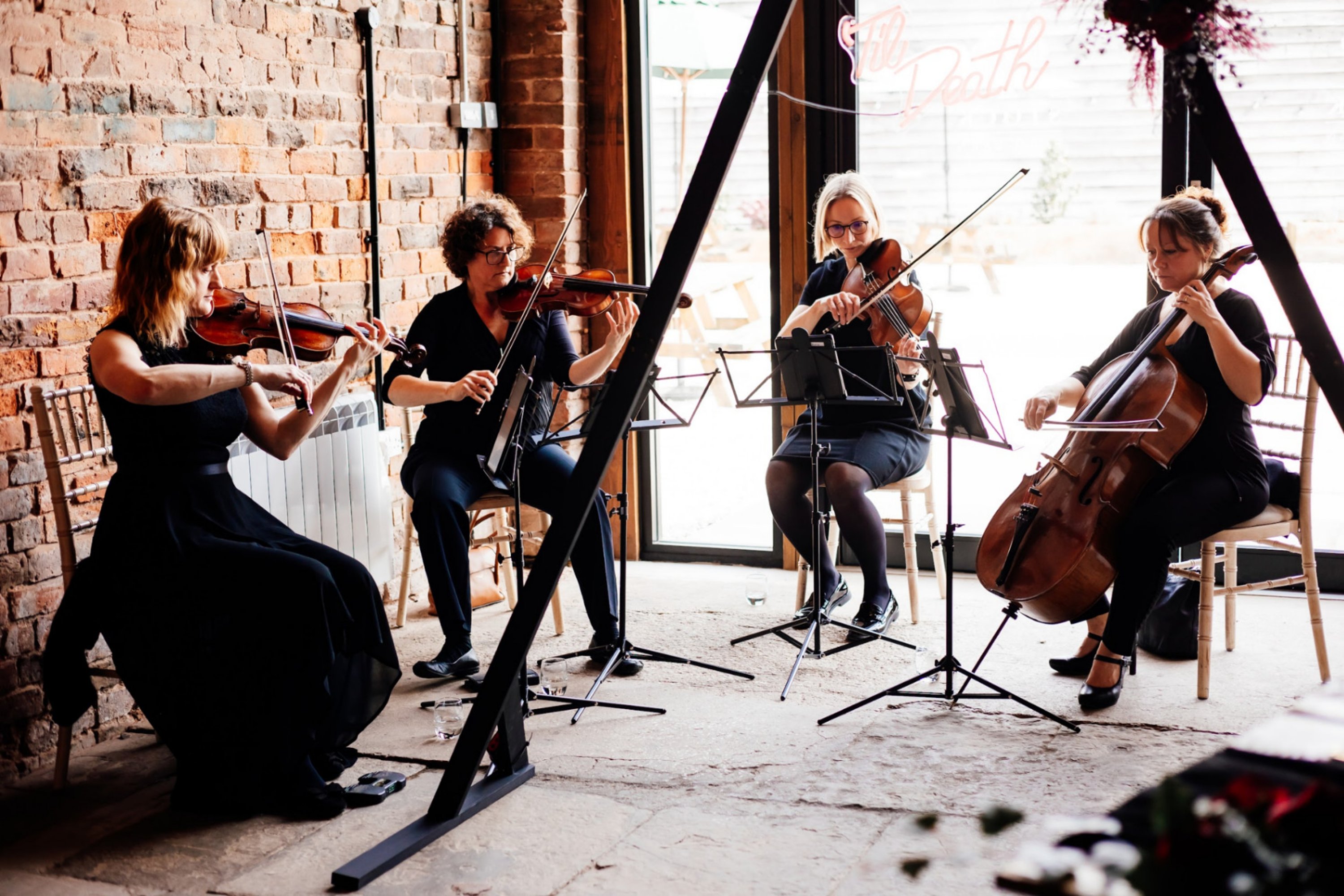 four members of a string quartet wearing black playing their violins and cellos in a brick room