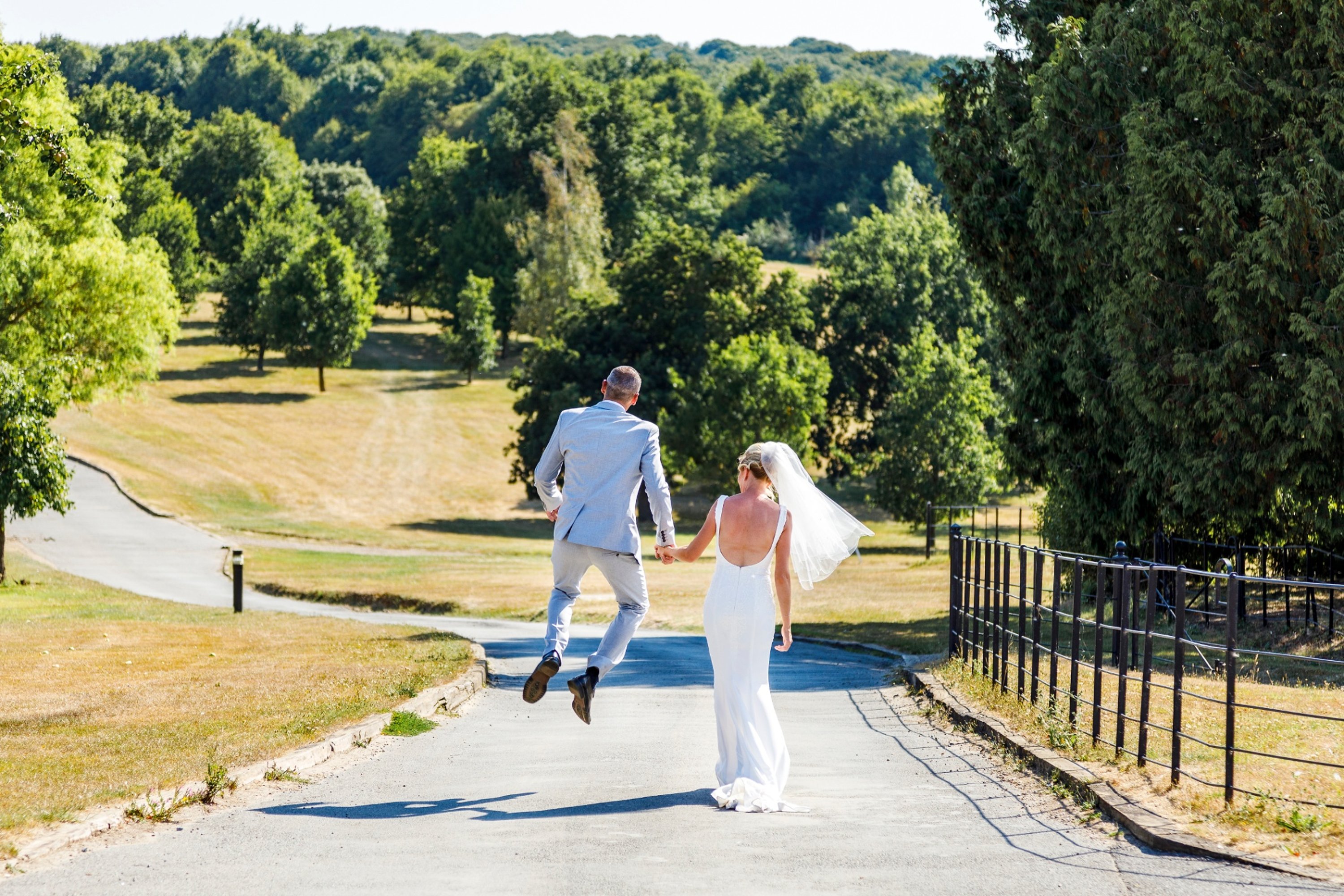 Groom in a pale blue suit holding hands with a bride as he walks down a driveway after getting married, leaping in the air with joy