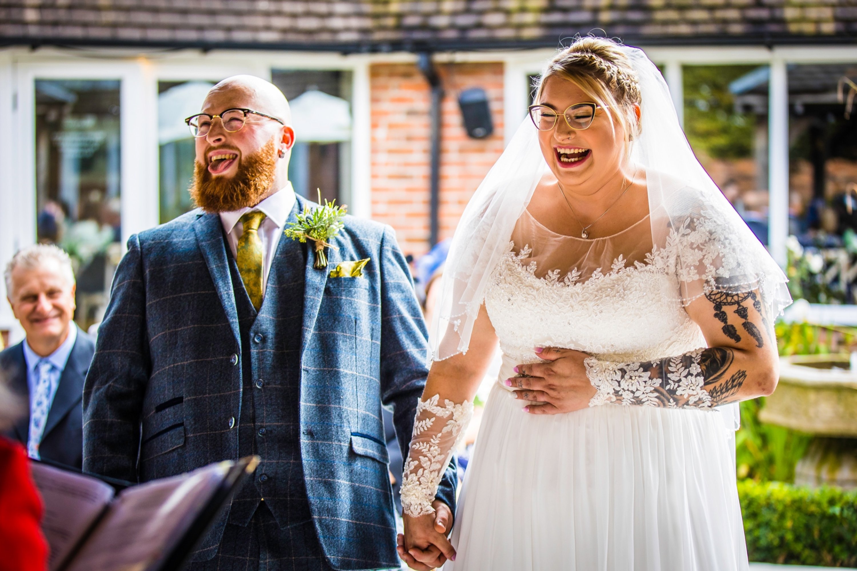 a bride laughing holding hands with her new husband, who is telling a joke in front of their home 