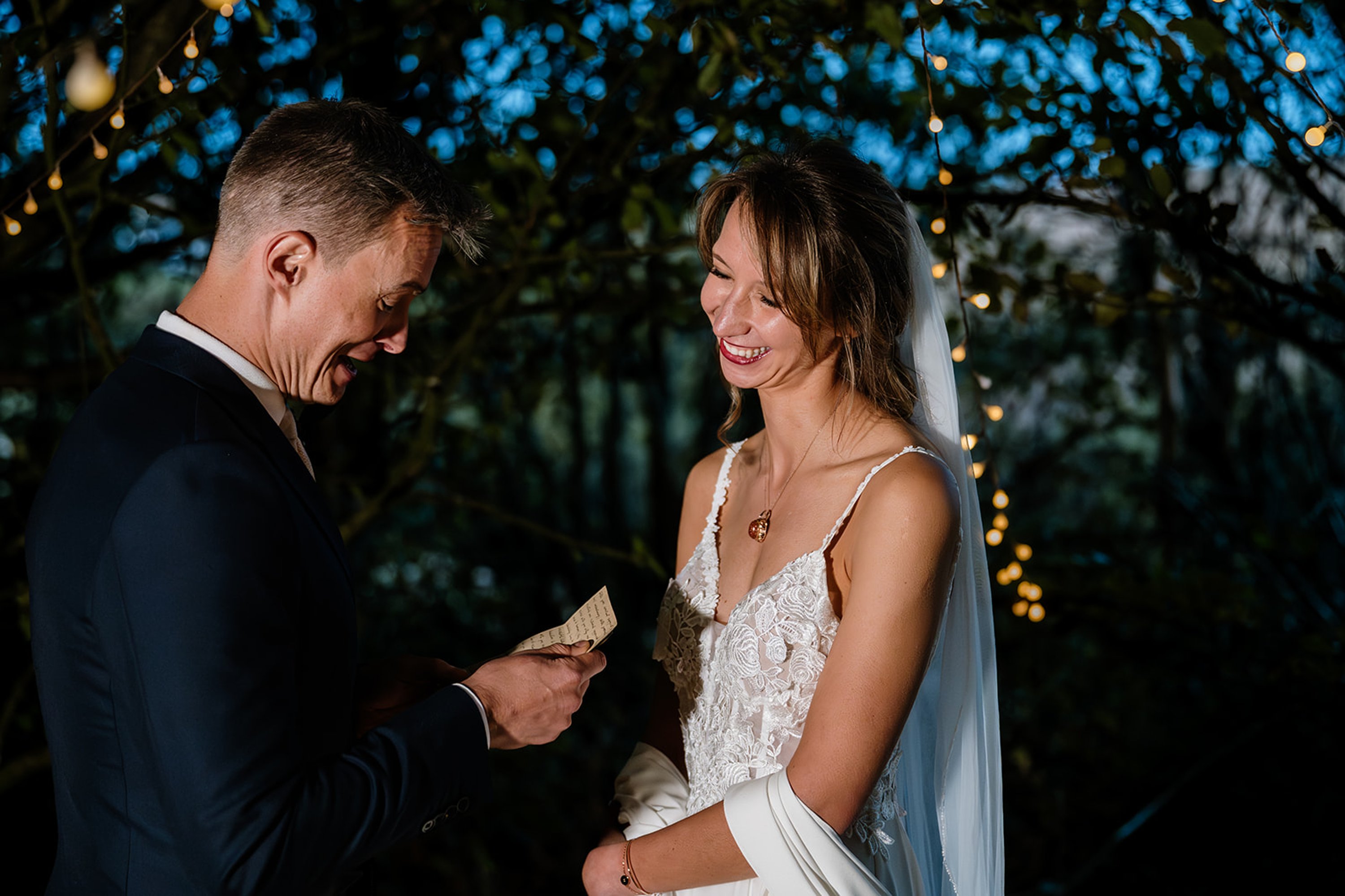 A bride laughing at a her groom reading his speech at twilight surrounded by fairy lights