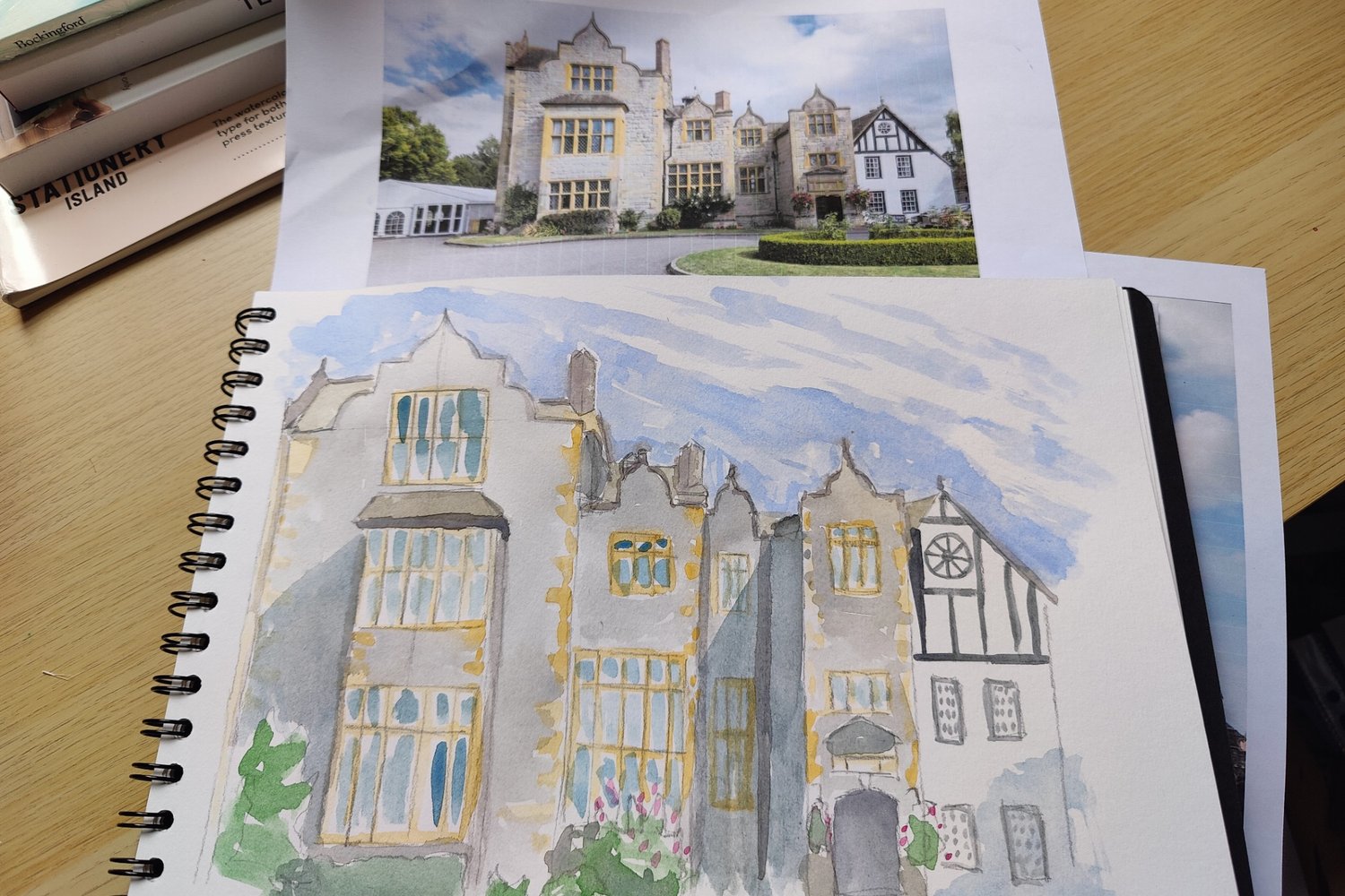 A large open sketchbook of a watercolour painting showing the outside of a wedding venue stately home