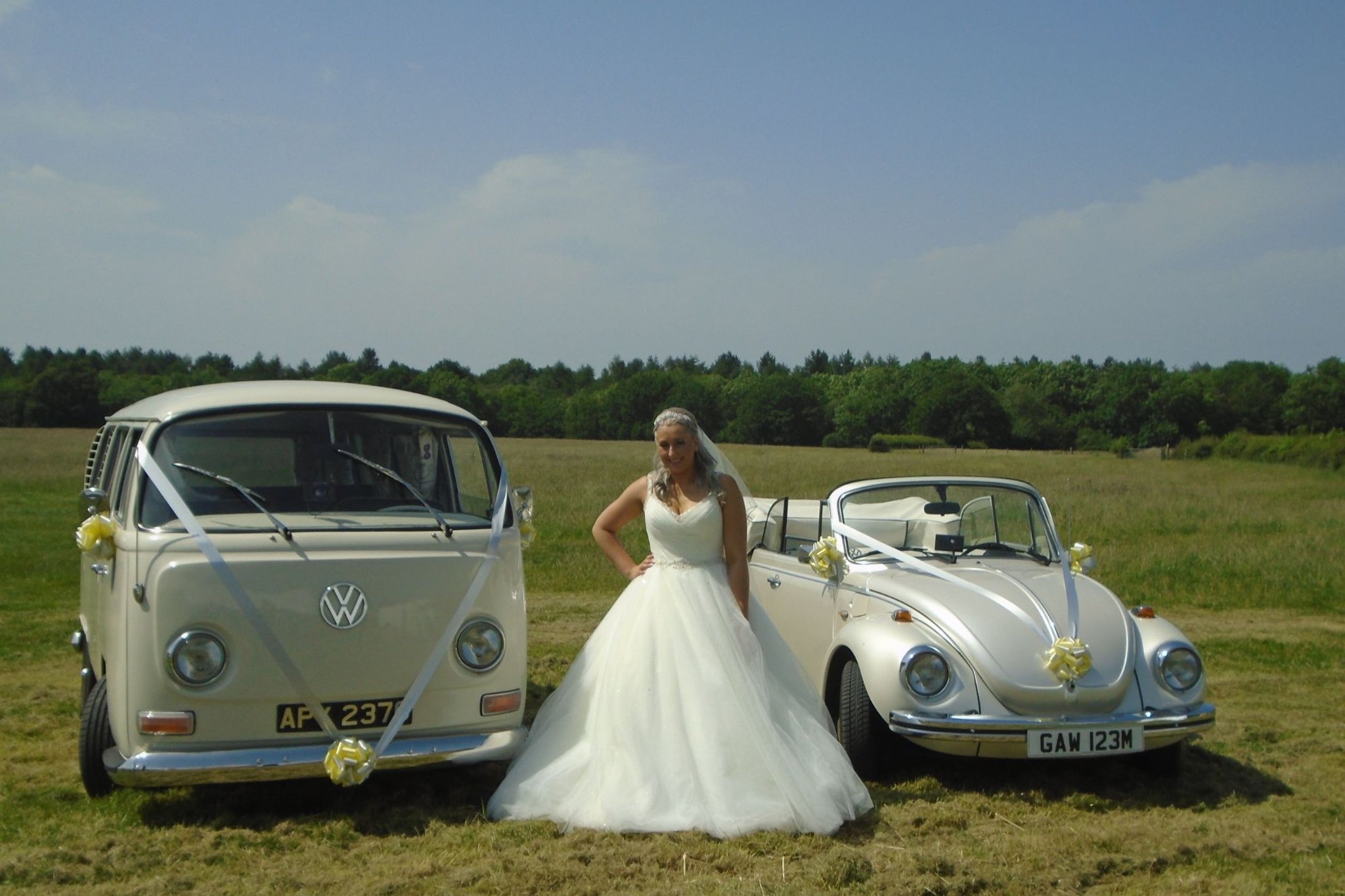 A bride posing next to a vintage VW camper van and a convertible Beetle decorated for weddings