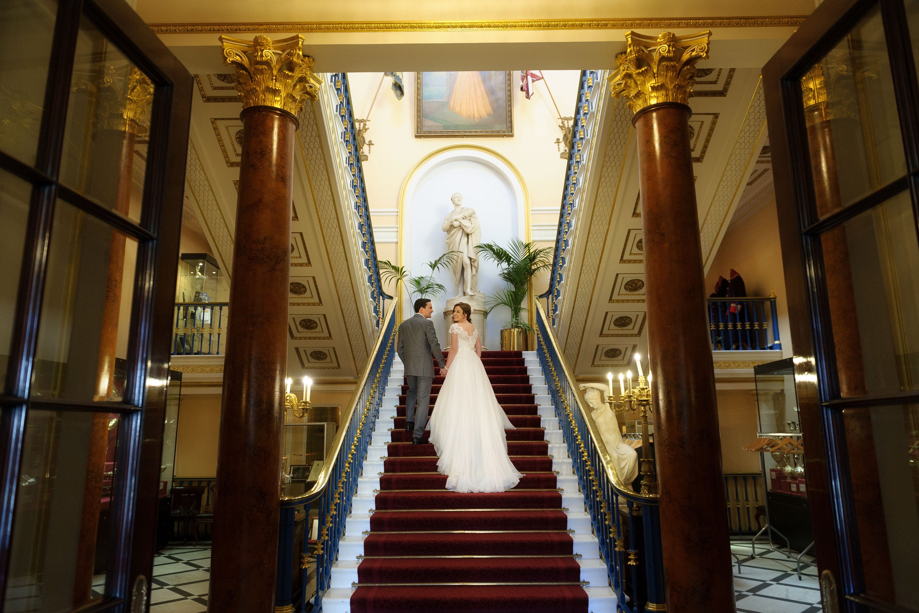 A bride and groom walking up the stairs at Liverpool Town Hall, in a grand gold and red drawing room